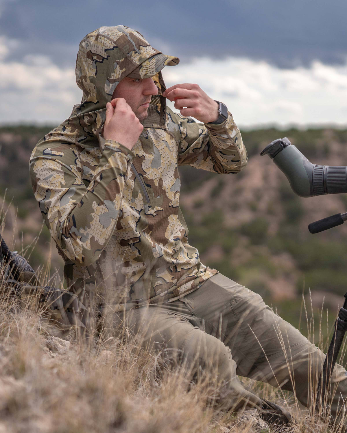 Hunter sitting on a hillside adjusting the hood of his Tiburon Hybrid Hoodie in Valo Camouflage