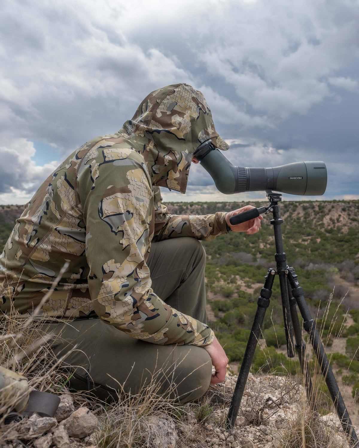 Side view of hunter crouched down, looking through his spotting scope, wearing Tiburon Hybrid Hoodie in Valo Camouflage