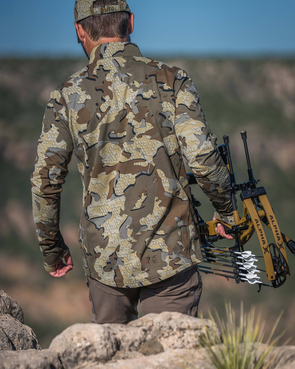 Back view of a hunter wearing Vital S3 Zip-T in Valo Camouflage carrying a yellow and black compound bow with arrows on a rocky outdoor hillside.