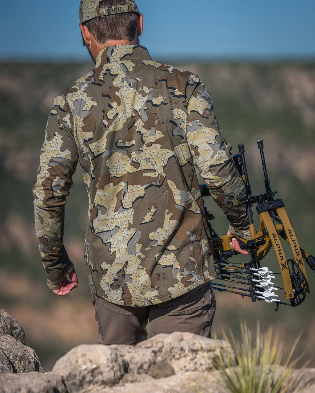 Back view of a hunter wearing Vital S3 Zip-T in Valo Camouflage carrying a yellow and black compound bow with arrows on a rocky outdoor hillside.