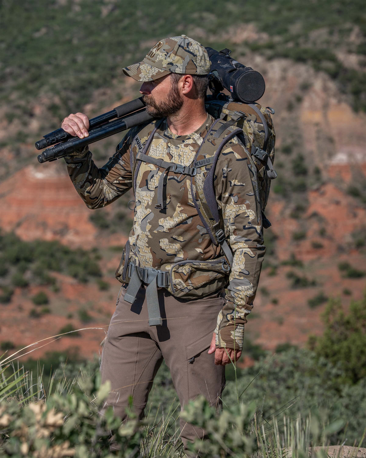 A man wearing a Vital S3 Long Sleeve Crew in Valo Camouflage walks through a rocky outdoor landscape while carrying a Valo Camouflage backpack and a black tripod over his shoulder.