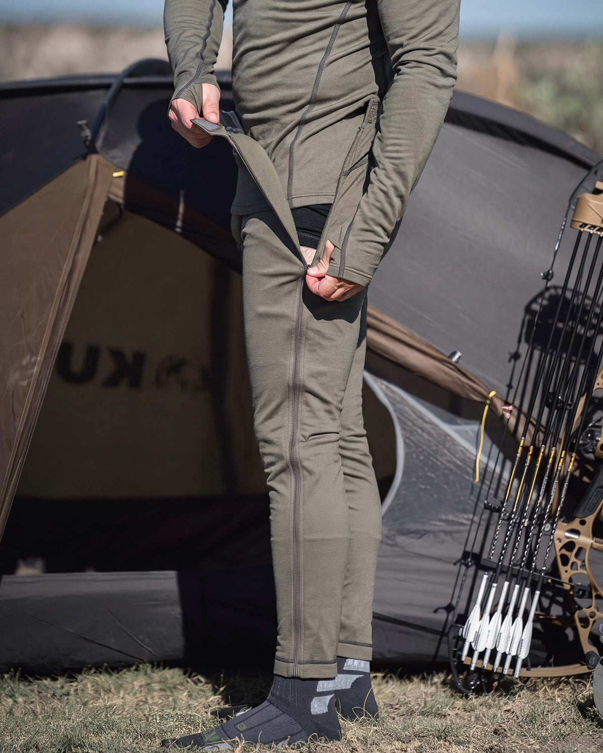 A person unzipping the size zipper on a Vital S3 Zip-Off Bottom in Ash Brown, stands outdoors in front of a tent, with a compound bow leaning against the tent on the right.