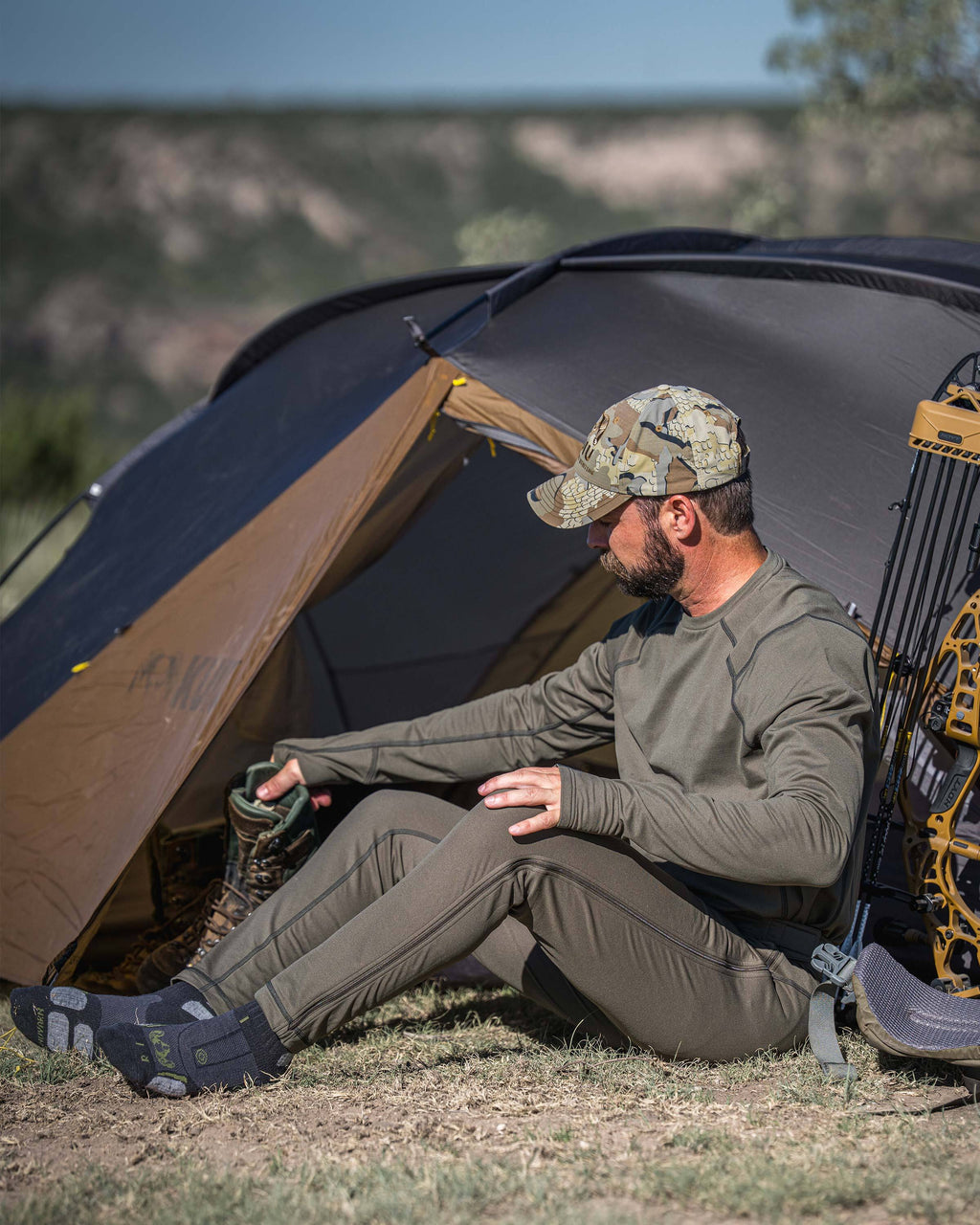 A man sits on the ground in front of a large camping tent, wearing a Vital S3 Zip-Off Bottom in Ash Brown.