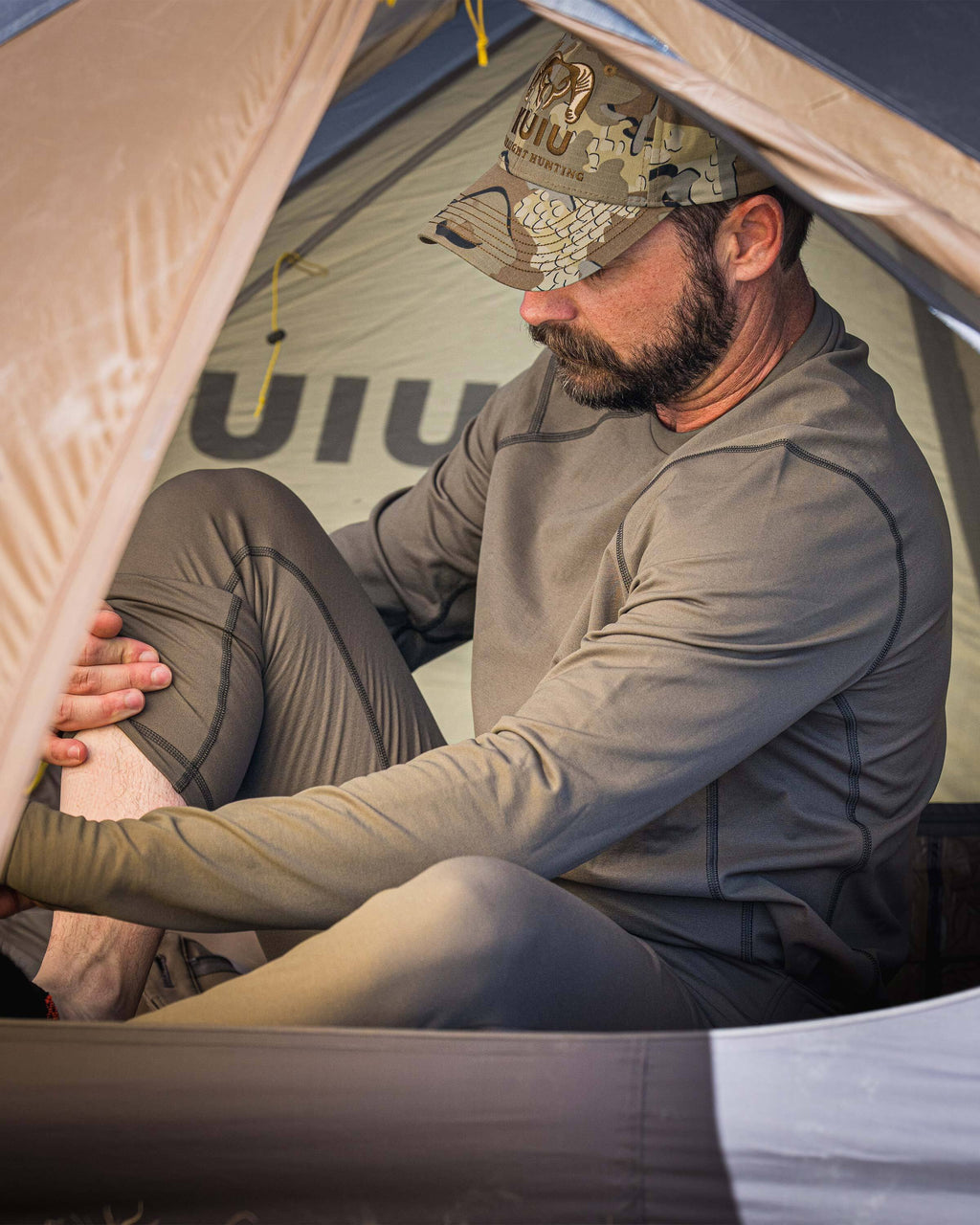 A man sitting inside a KUIU tent wearing a Vital S3 Long Sleeve Crew in Ash Brown and matching Vital S3 Zip-Off Bottom.