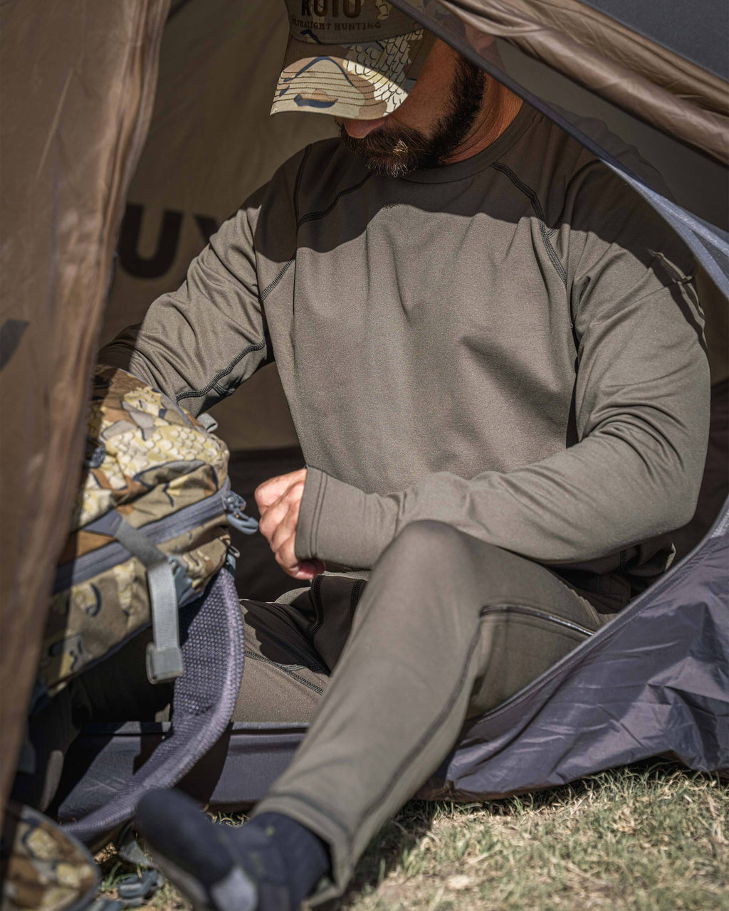 A man sitting inside a KUIU tent wearing a Vital S3 Long Sleeve Crew in Ash Brown and matching Vital S3 Zip-Off Bottom.