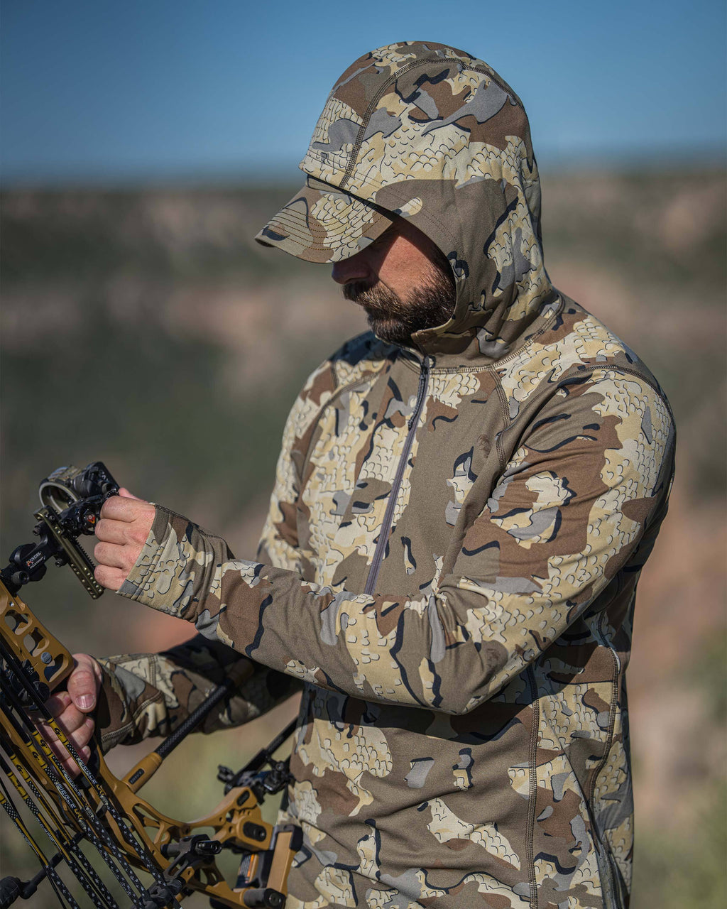 A man wearing a Vital S3 Zip-T Hoodie in Valo Camouflage holding a bow in an outdoor landscape setting.