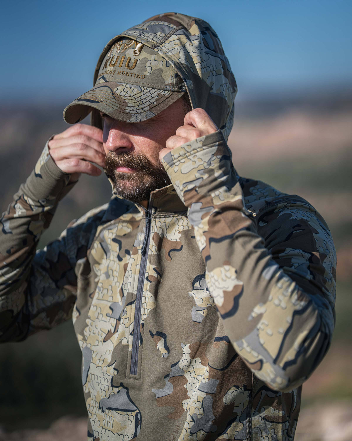 A man wearing a Vital S3 Zip-T Hoodie in Valo Camouflage adjusting his hoodie in an outdoor setting.