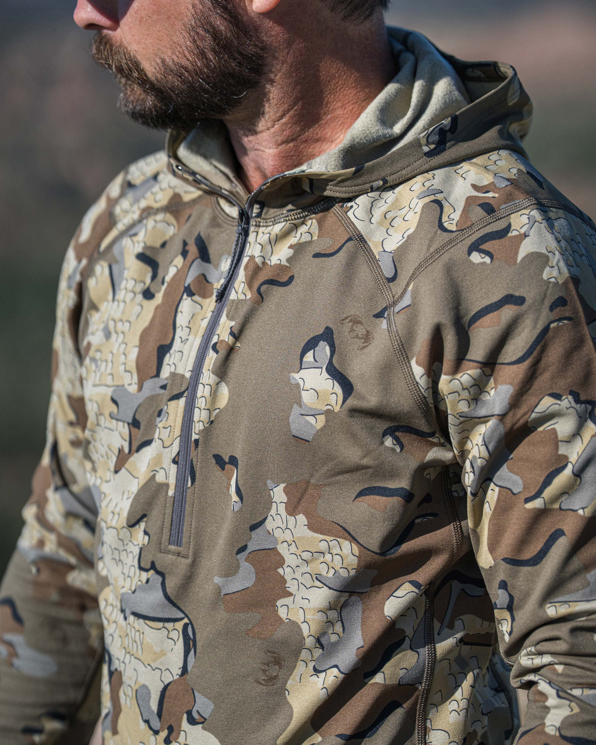 A man in profile wearing a Vital S3 Zip-T Hoodie in Valo Camouflage in outdoor setting with a blurred natural background.
