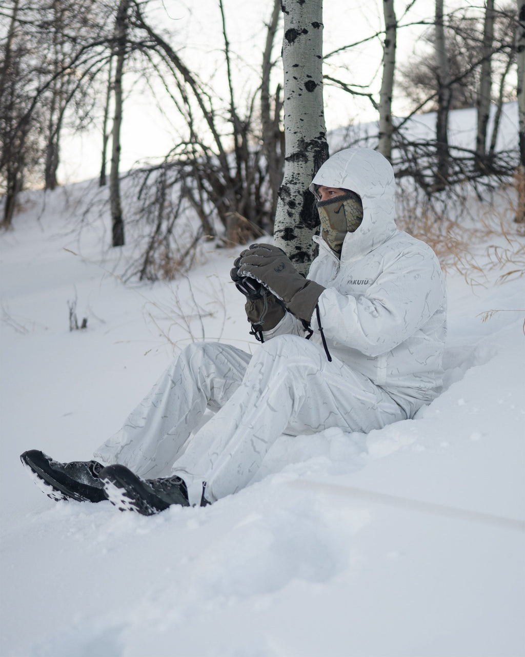Hunter wearing Snowfall Overwhites Set, sits on snow-covered ground holding a binocular 