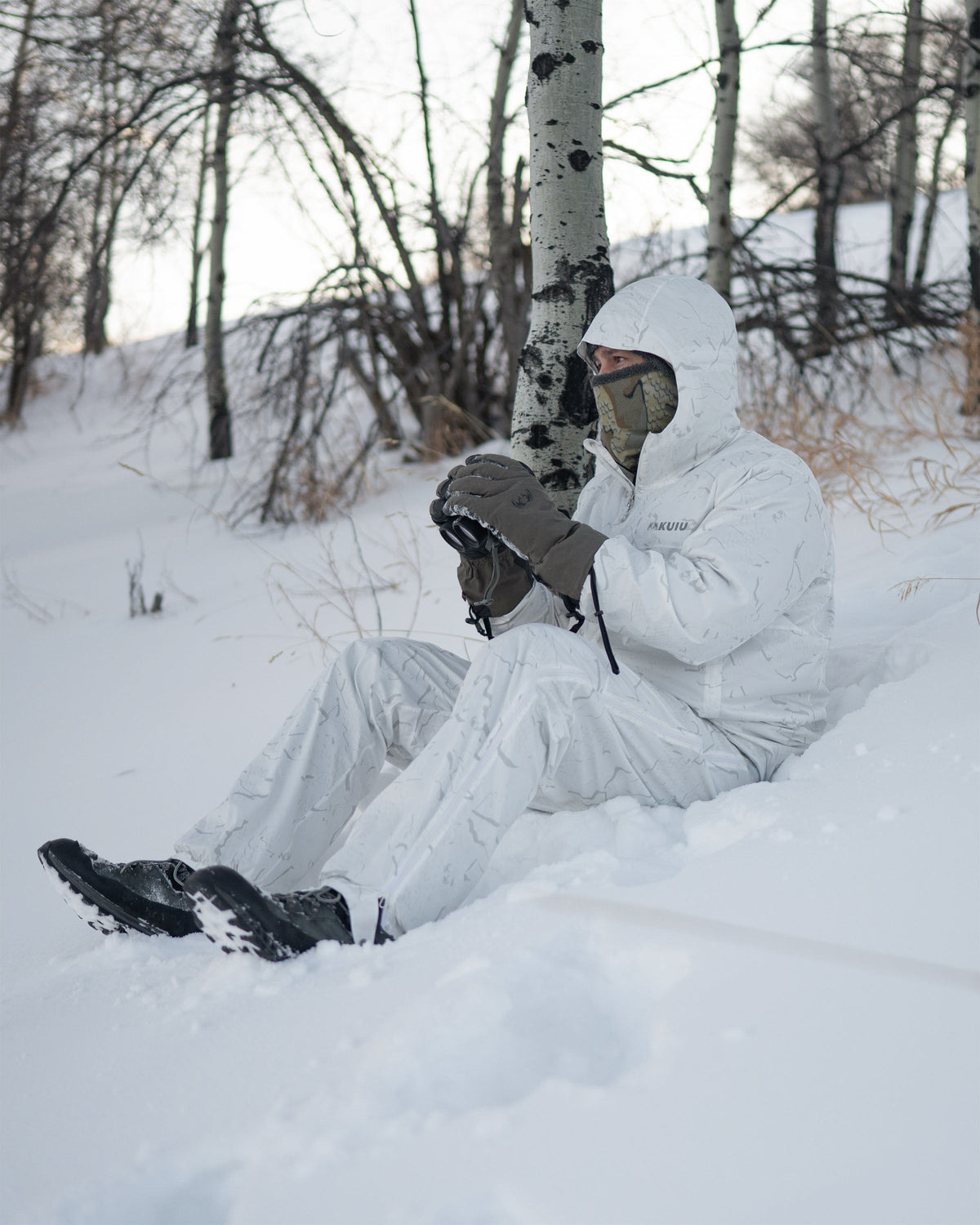 Hunter wearing Snowfall Overwhites Set, sits on snow-covered ground holding a binocular 