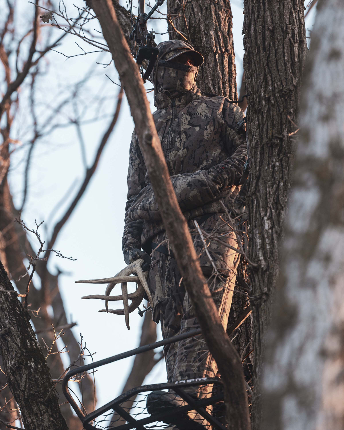 Whitetail hunter stands in a tree stand among tree trunks, wearing Proximity Insulated Pant in Veros Camouflage, holding antlers in a wooded outdoor setting.