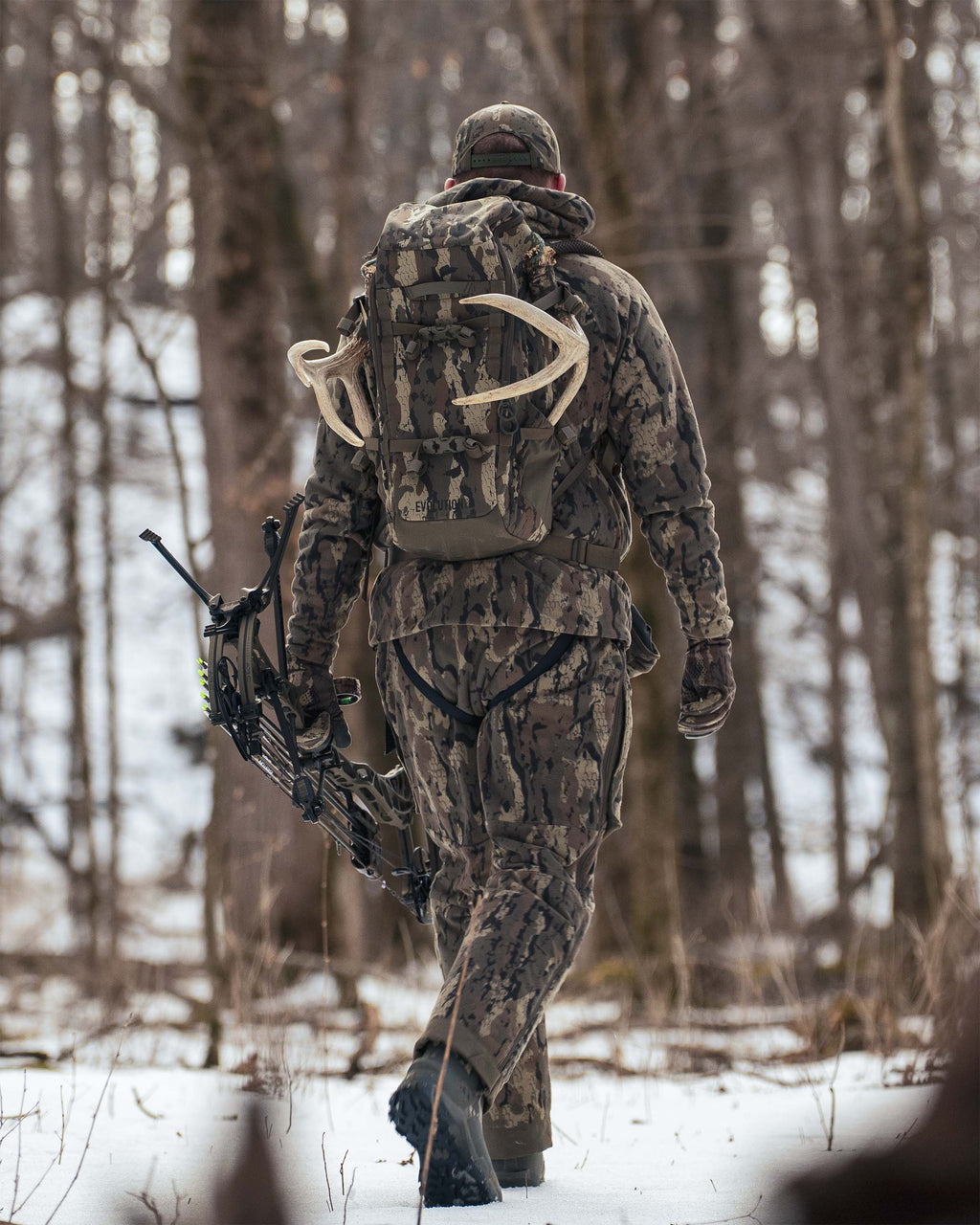 Hunter walking through a snowy forest, seen from behind wearing Proximity Insulated Pant in Veros Camouflage, carrying a Evolution 1800 backpack in Veros Camouflage.