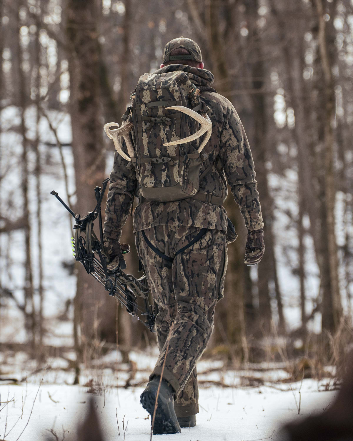 Hunter walking through a snowy forest, seen from behind wearing Proximity Insulated Pant in Veros Camouflage, carrying a Evolution 1800 backpack in Veros Camouflage.
