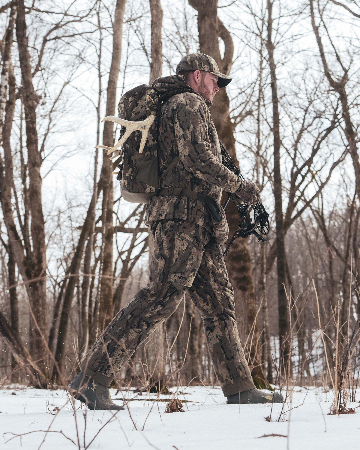 Hunter wearing Proximity Insulated Pant in Veros Camouflage walking through a snowy winter forest with bare trees. He is carrying a Evolution 1800 backpack in Veros Camouflage with a deer antler attachment and holding a black compound bow.
