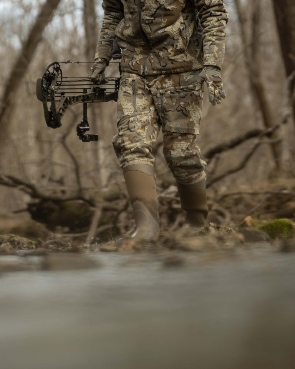 Front of Hunter Holding Bow Standing Next to Creek Wearing Proximity Insulated Pants in Valo Off-Shade Camouflage