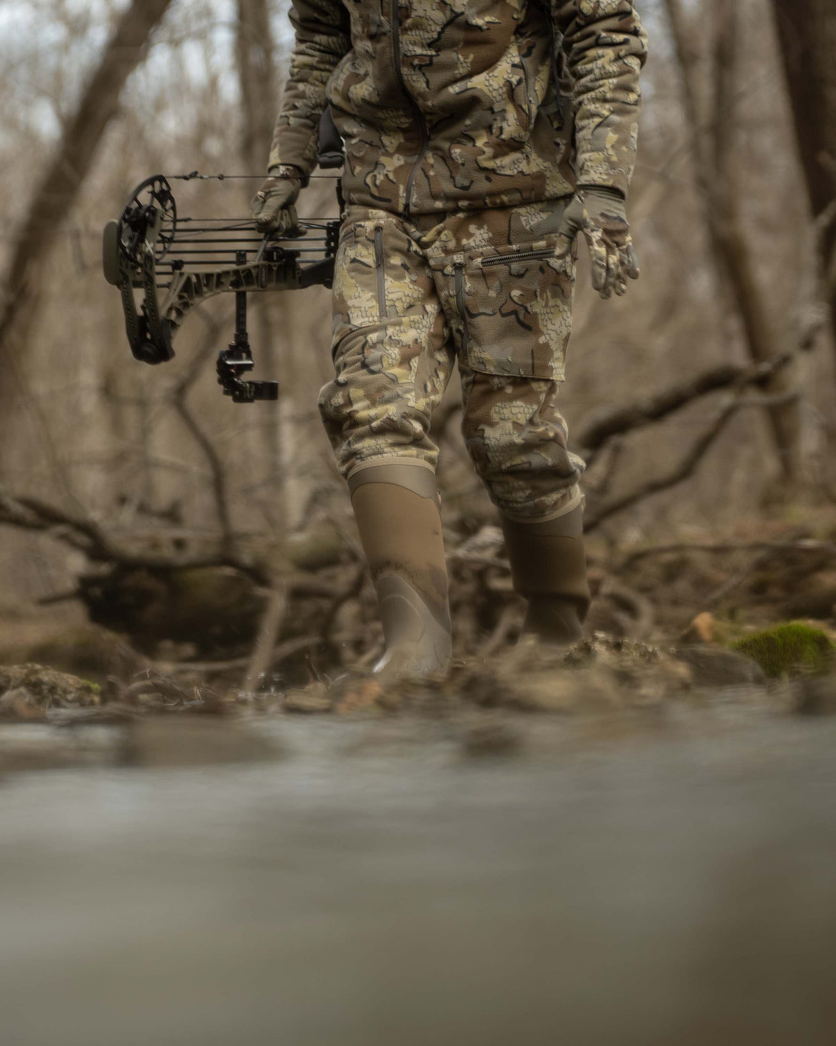 Front of Hunter Holding Bow Standing Next to Creek Wearing Proximity Insulated Pants in Valo Off-Shade Camouflage