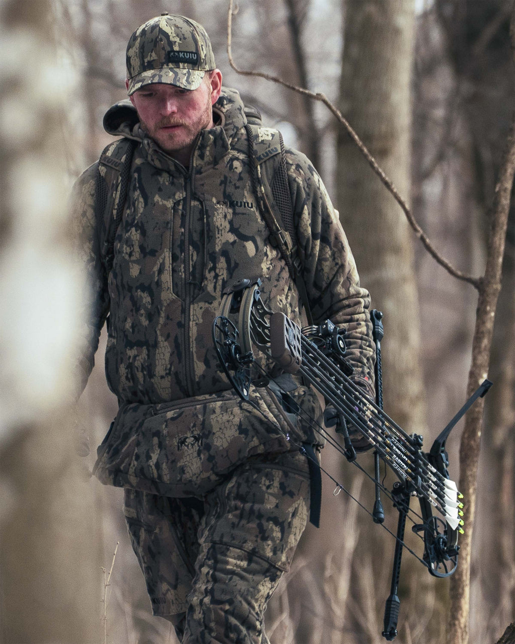 Hunter wearing Proximity Hooded Jacket in Veros camouflage and insulated pants, walks through a forest while carrying a compound bow on his back.