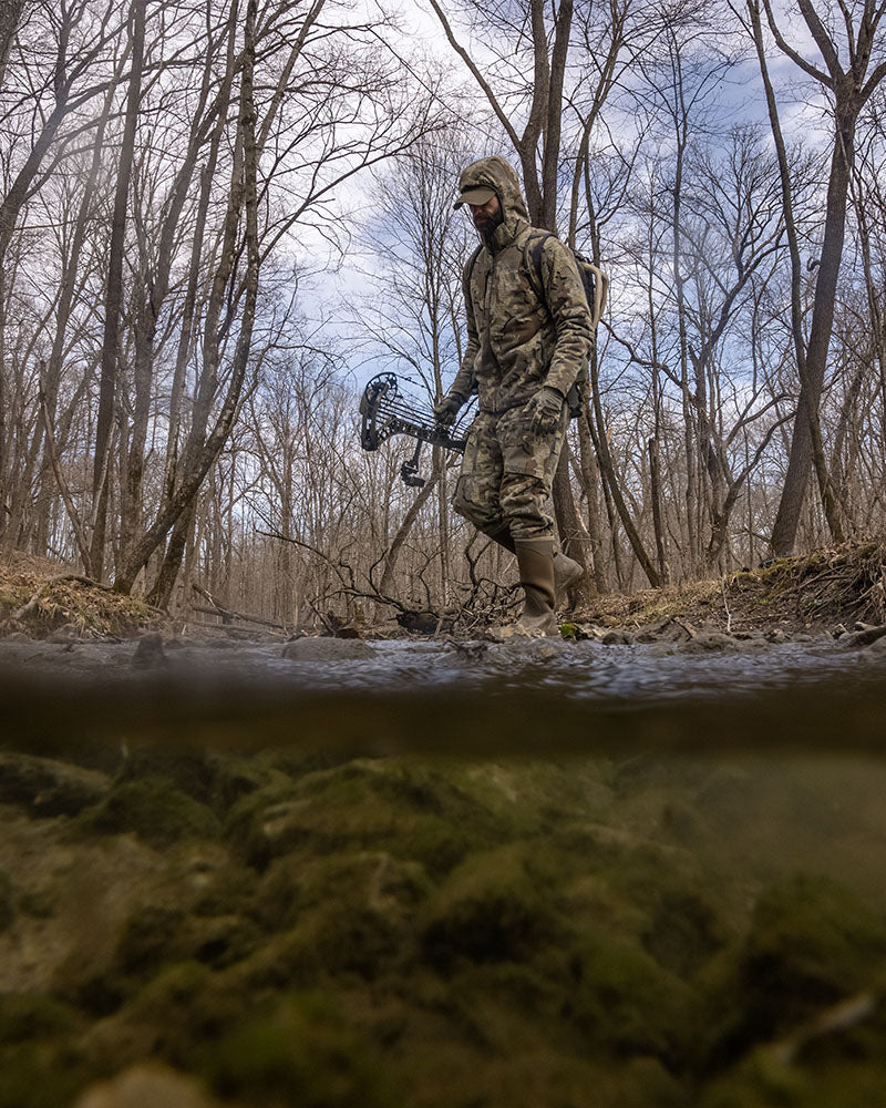 Side of Hunter Holding Bow Walking Through Creek Wearing Proximity Hooded Insulated Jacket in Valo Camouflage