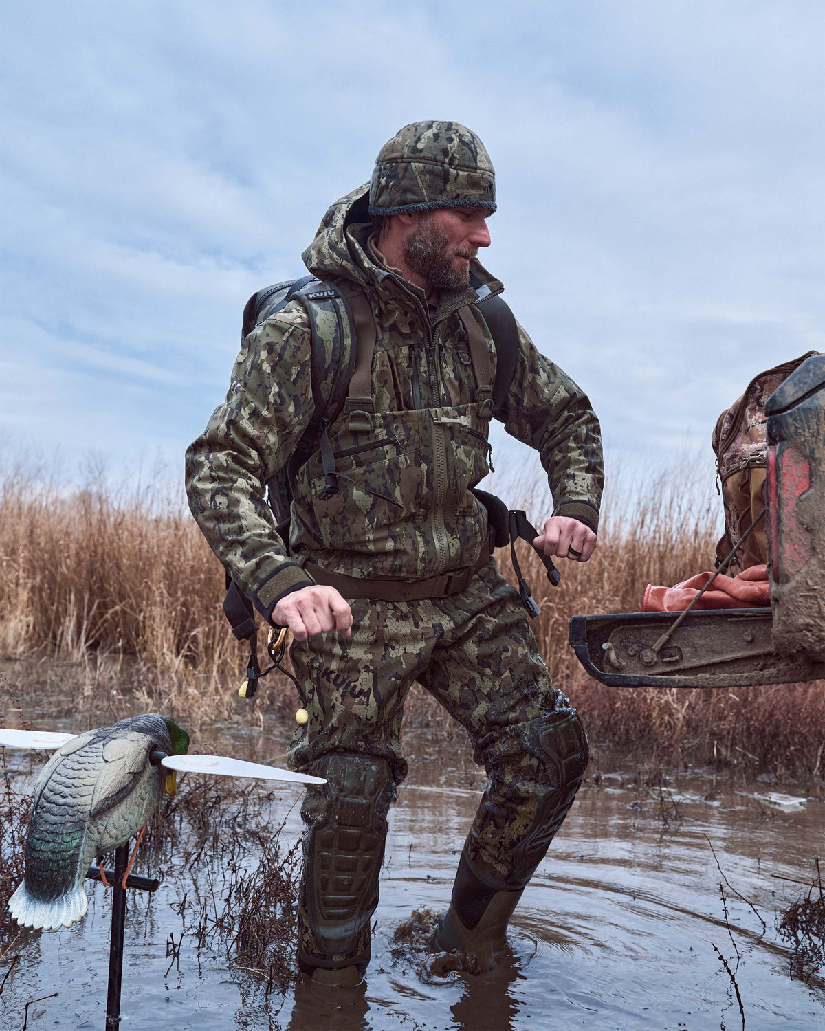 Hunter wearing Proximity Beanie in Veros Camouflage stands in shallow water, holding a bow with a duck on it. 