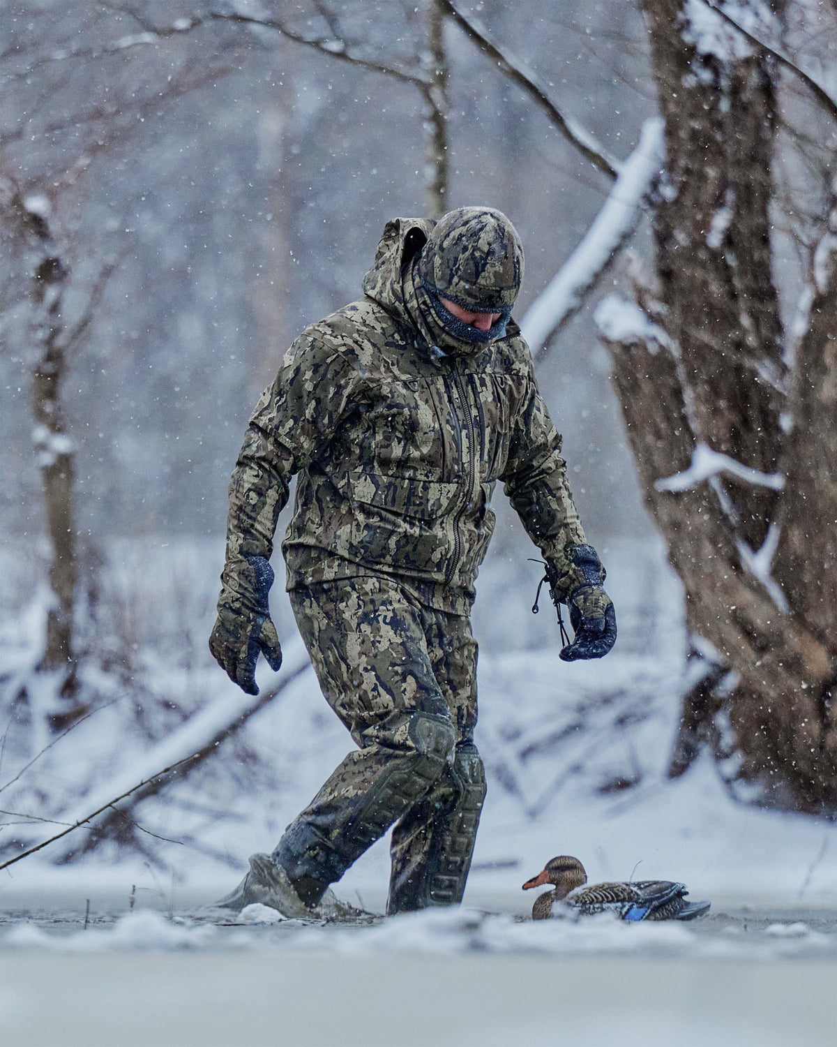Hunter in full Veros camouflage clothing, Proximity neck gaiter, beanie, gloves, and wader walks through snow in a wooded outdoor setting, with a duck visible on the ground nearby.