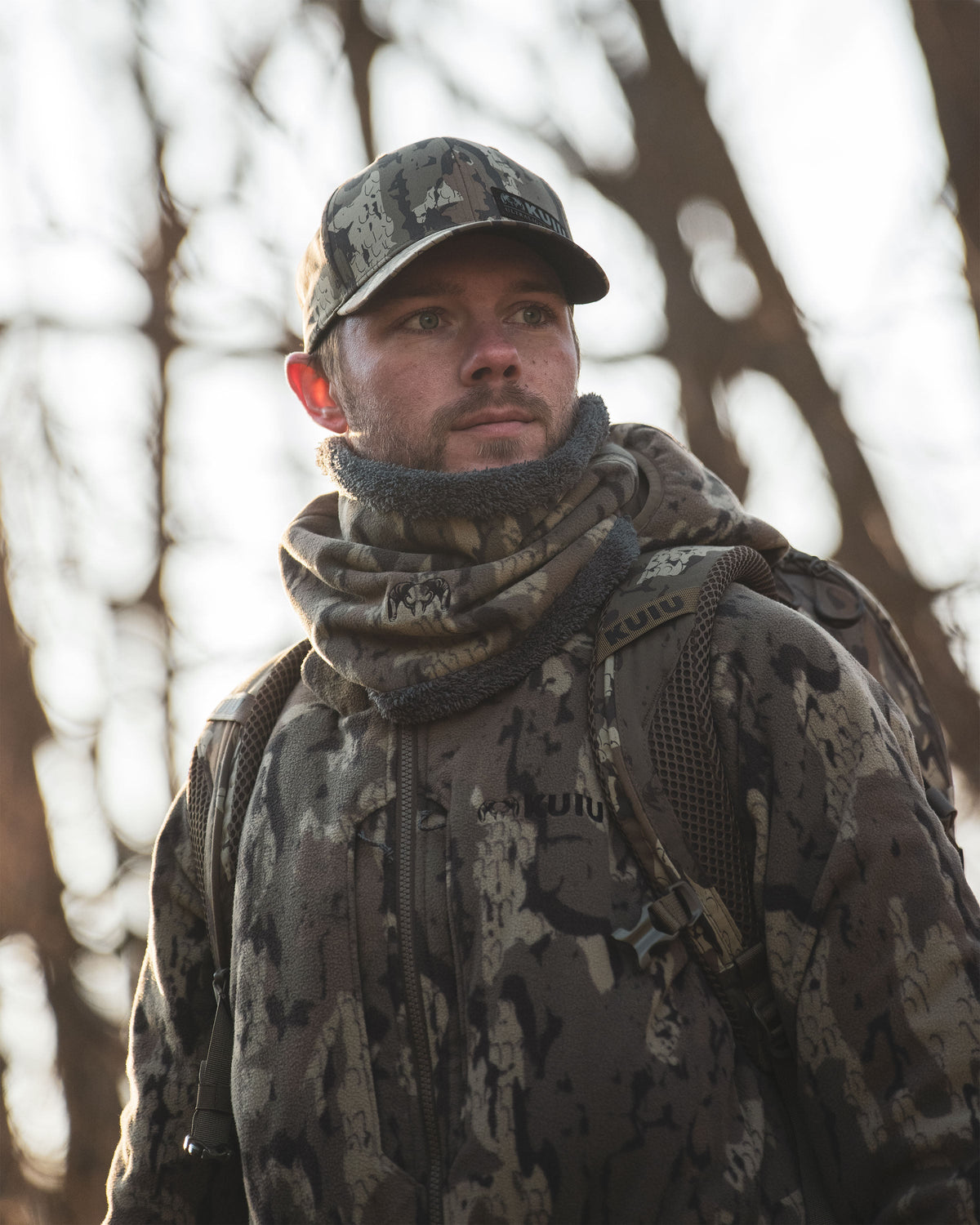 Hunter wearing Proximity Neck Gaiter in Veros Camouflage standing outdoors in a wooded area with bare trees in the background.