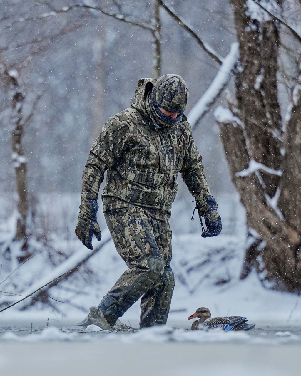 Hunter in full Veros camouflage clothing, Proximity neck gaiter, beanie, gloves, and wader walks through snow in a wooded outdoor setting, with a duck visible on the ground nearby.