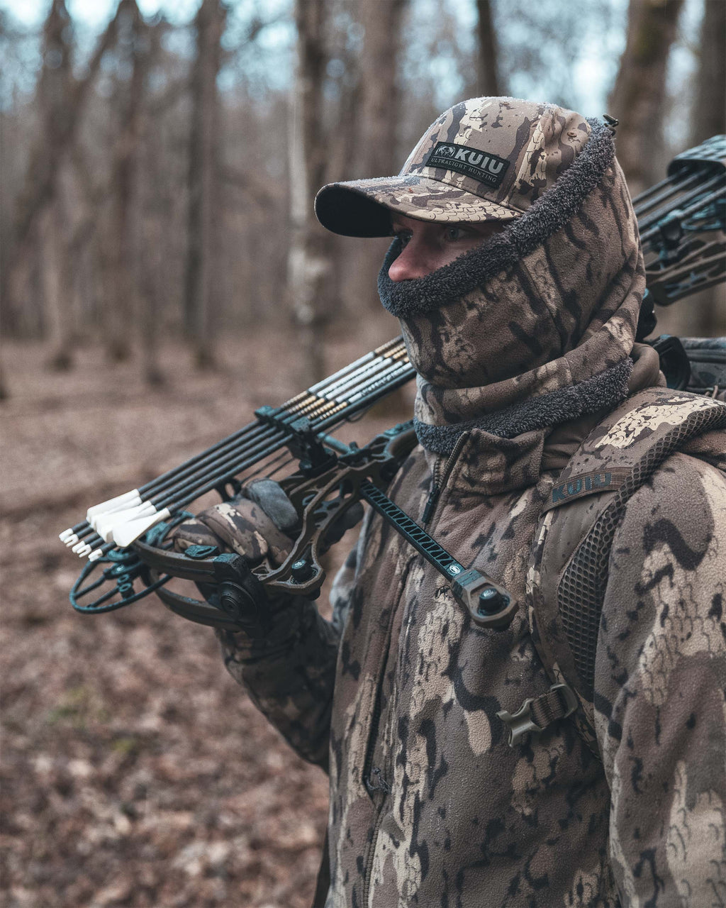 Hunter wearing Proximity Neck Gaiter in Veros camouflage stands in a wooded forest setting, holding a compound bow. 