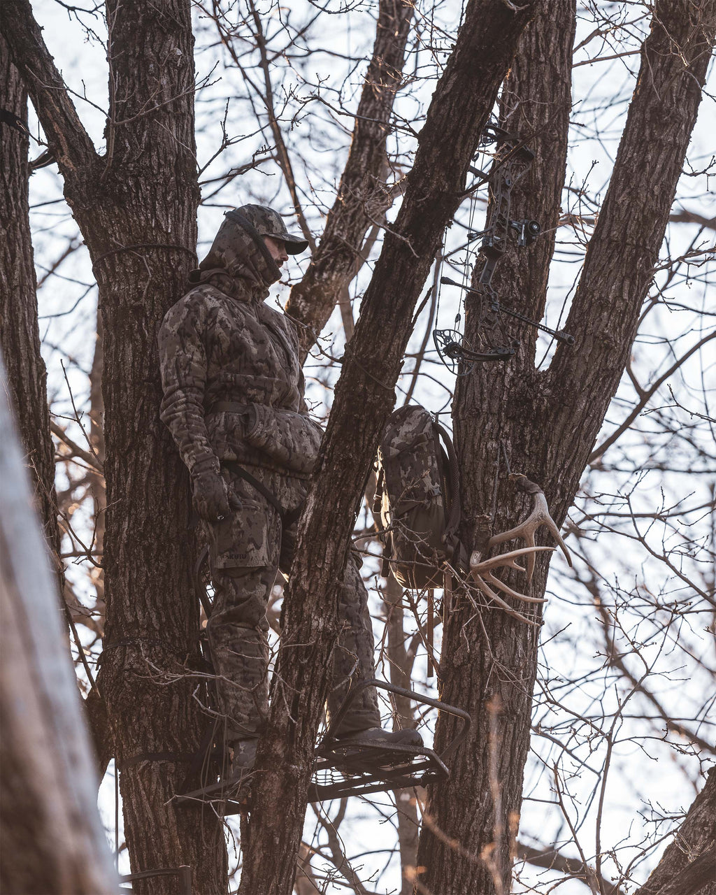 Hunter in full KUIU Veros camouflage hunting gear stands in a tree stand among bare trees, with a Evolution 1800 in Veros camouflage mounted on a branch nearby.