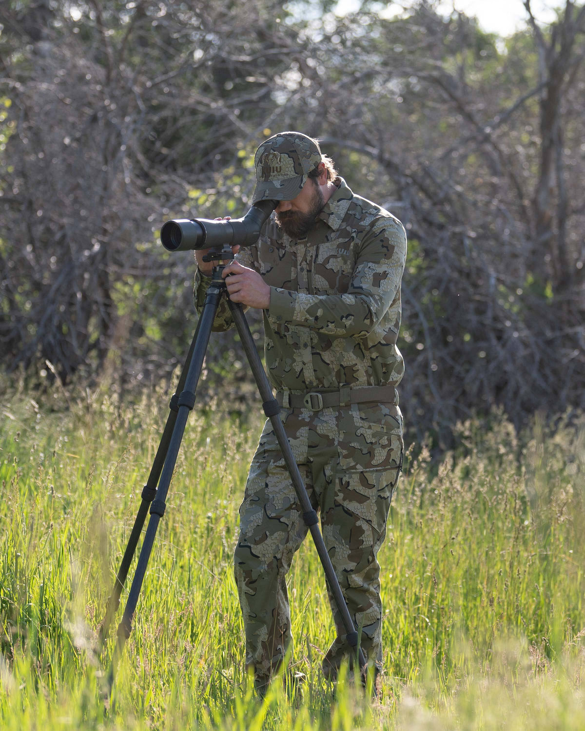 Hunter looking through a spotting scope in a grassy field, wearing Encounter LT Pant in Valo Camouflage