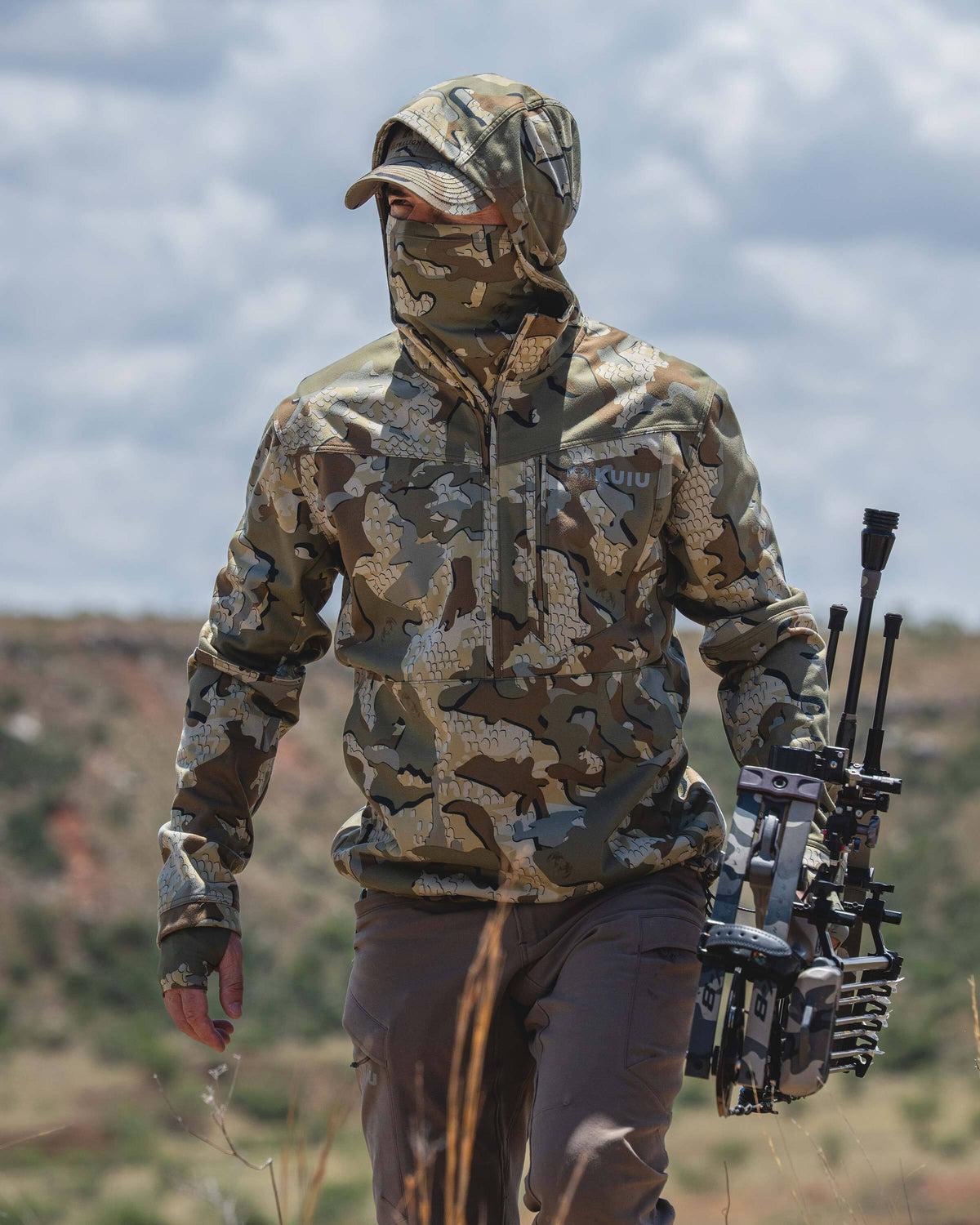 A person wearing a Drawpoint Hoodie in Valo camouflage walks outdoors while carrying a compound bow. The background shows a blurred natural landscape under a cloudy sky.