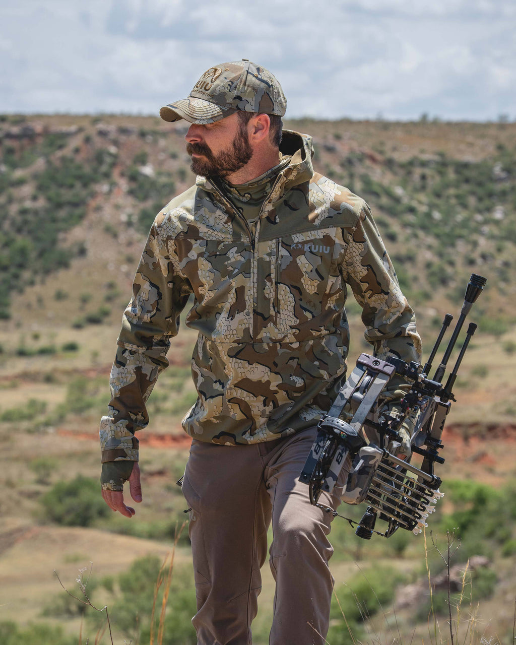 A man wearing a Drawpoint Hoodie in Valo camouflage walks through an outdoor desert landscape while carrying a compound bow.