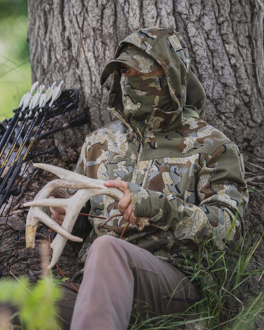 Hunter wearing Drawpoint Hoodie in Valo camouflage hooded with face covering sits under a tree holding a set of antlers.