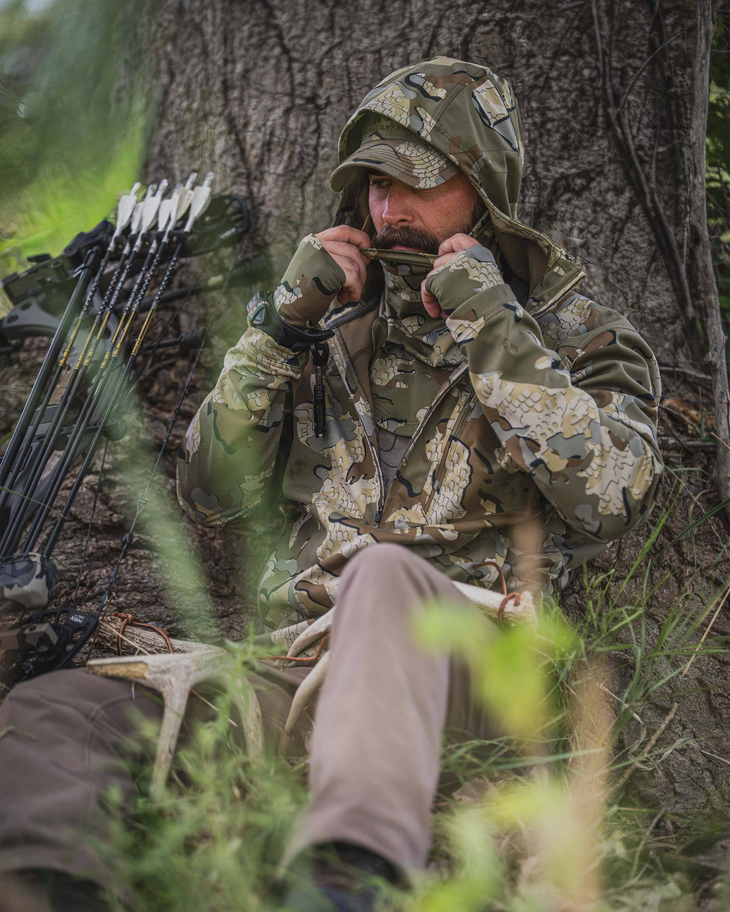 A man wearing Drawpoint Hoodie in Valo camouflage sits outdoors against a large tree, pulling the built-in face covering over his mouth. Several arrows are visible leaning against the tree on the left, with grass and foliage in the foreground.