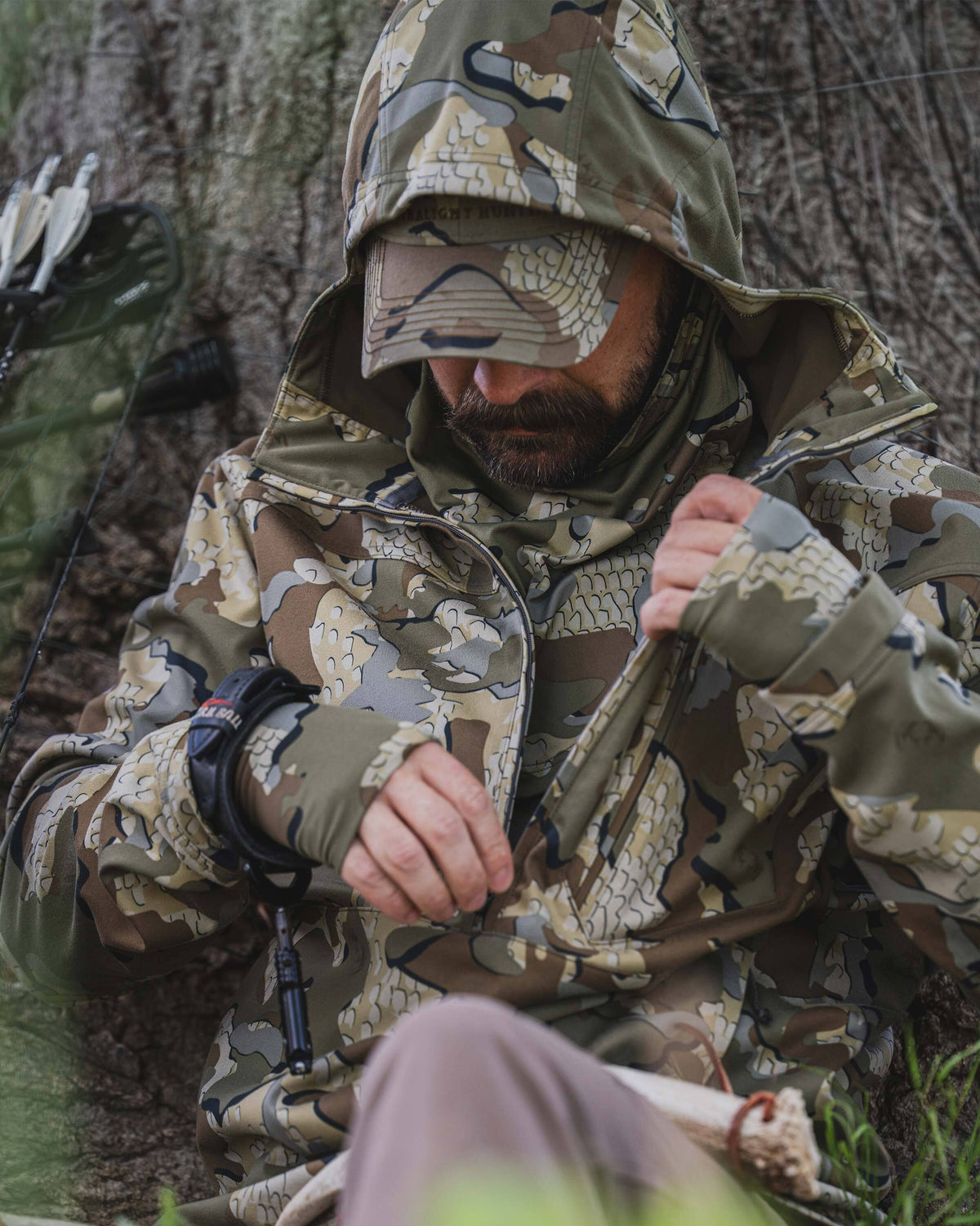 A man wearing a Drawpoint Hoodie in Valo camouflage sits outdoors against a tree trunk, adjusting the front zipper.