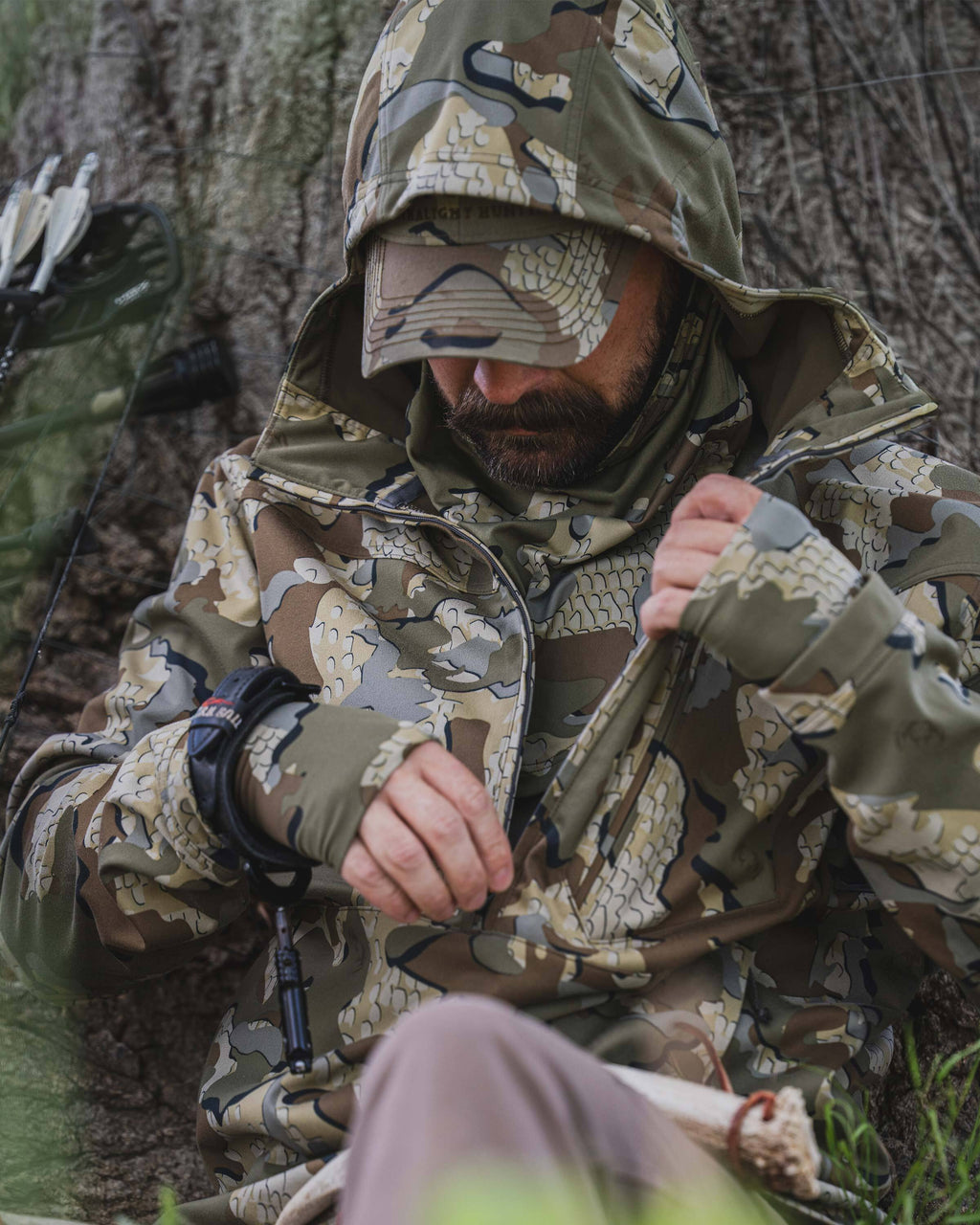 A man wearing a Drawpoint Hoodie in Valo camouflage sits outdoors against a tree trunk, adjusting the front zipper.