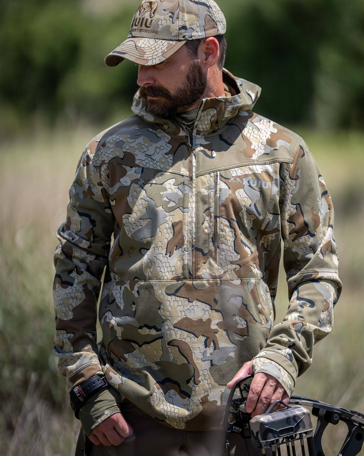 A man wearing a Drawpoint Hoodie in Valo camouflage and matching camouflage cap, standing outdoors in a grassy field.