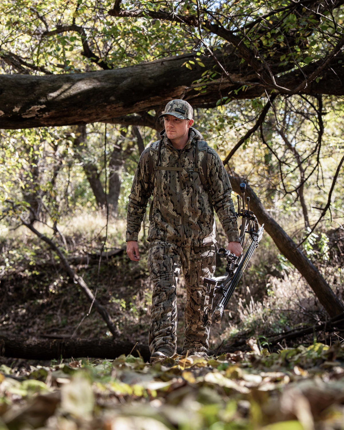 Hunter in full Veros camouflage hunting apparel walks through a wooded forest carrying a compound bow.