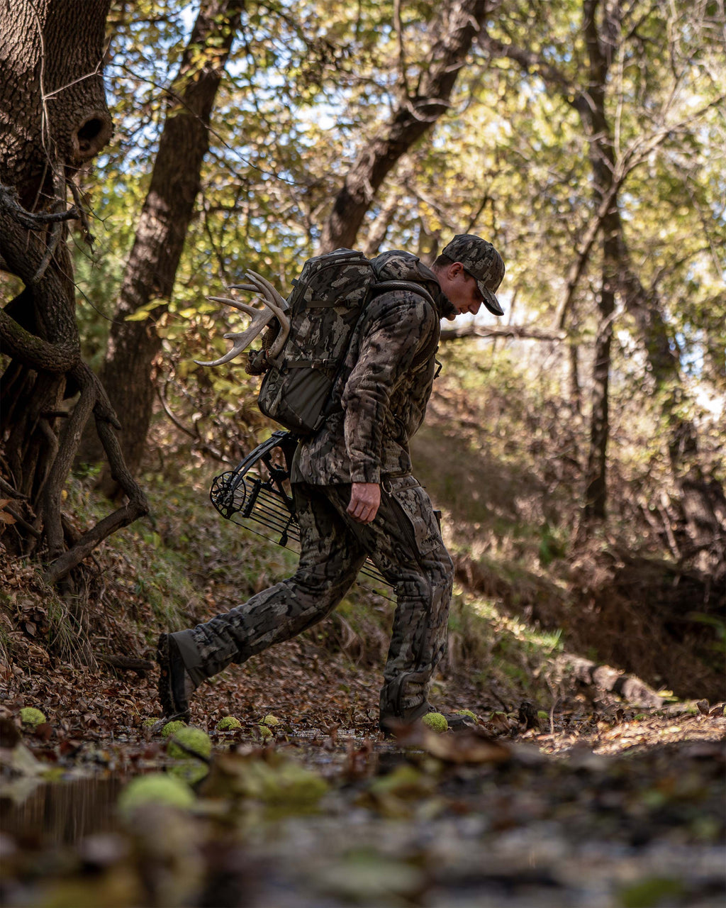 Hunter in full Veros camouflage hunting apparel walks through a wooded stream carrying a large camouflage backpack with a mounted bow visible behind him.