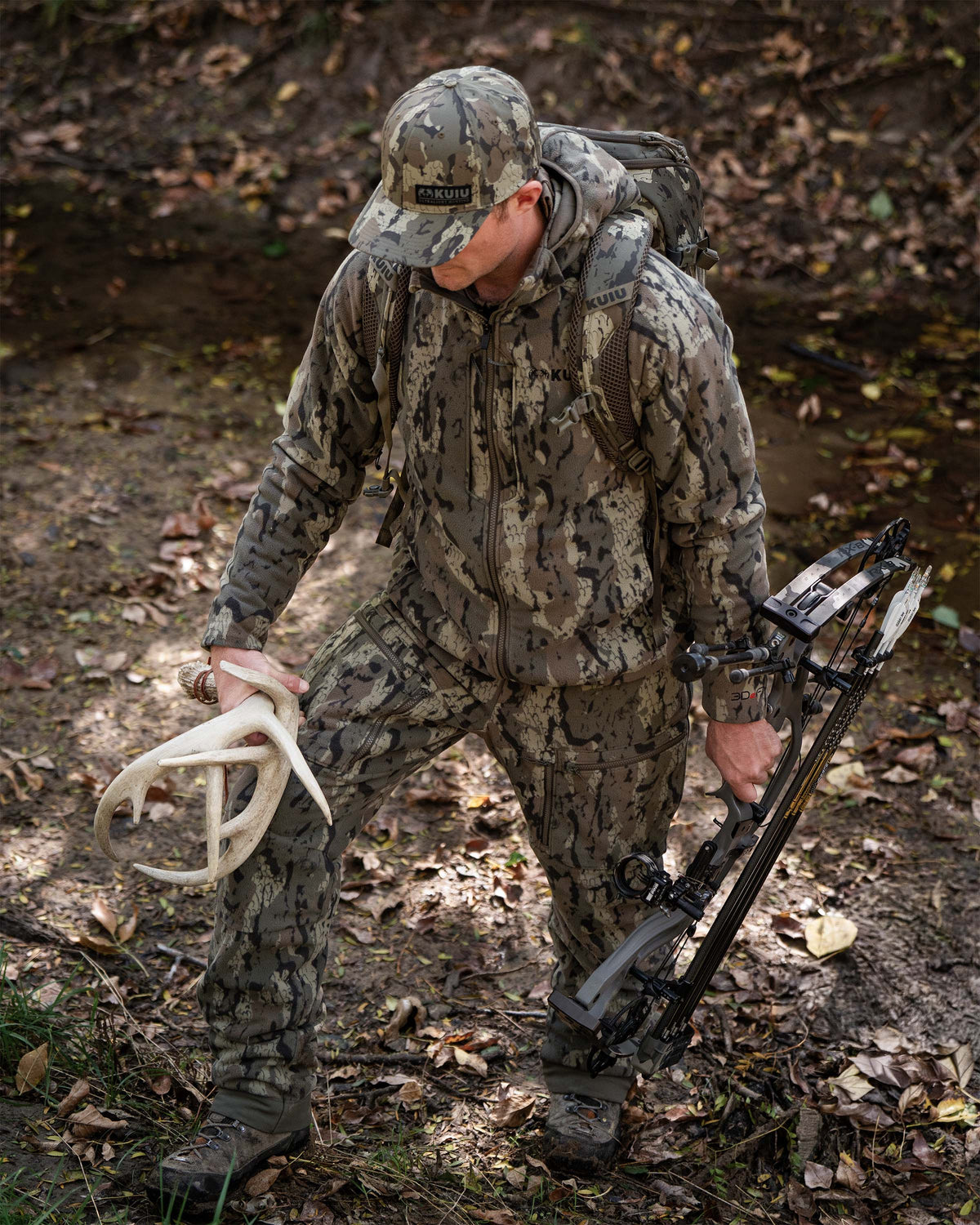 Hunter in full Veros camouflage hunting jacket, pants, and hat stands in a wooded outdoor setting holding a compound bow and a set of antlers.