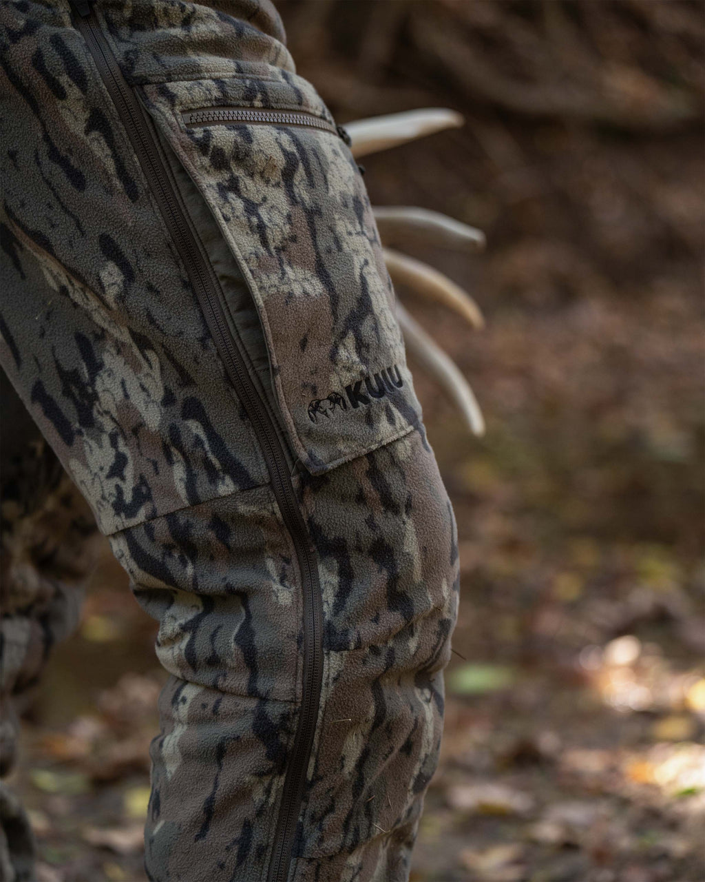 Close-up of Veros camouflage Proximity Insulated Bib with a vertical leg zipper and a visible KUIU logo, worn outdoors on a forest floor.