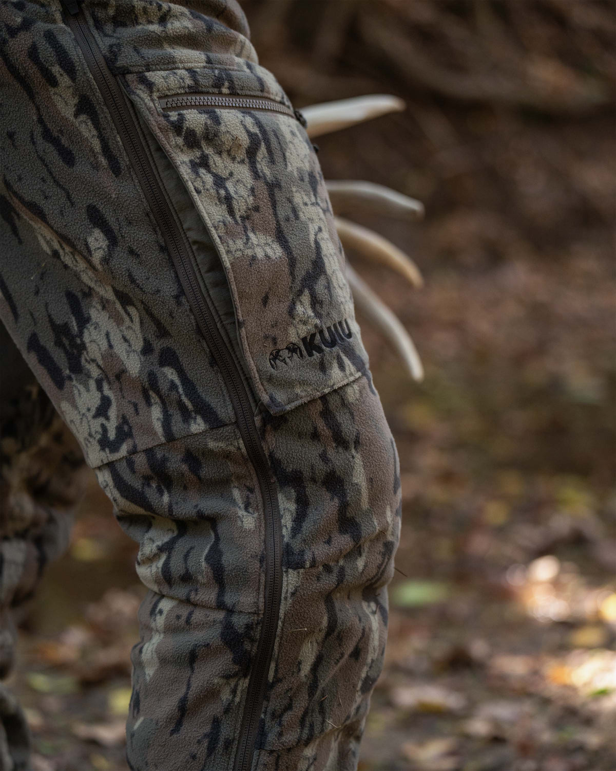 Close-up of Veros camouflage Proximity Insulated Bib with a vertical leg zipper and a visible KUIU logo, worn outdoors on a forest floor.
