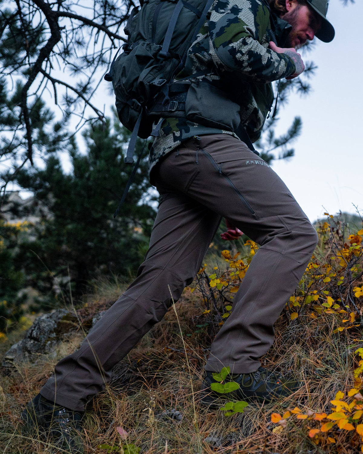 Side of Hunter Hiking Up Hill Wearing Attack Pants in Major Brown Color Highlighting YKK Zippered Hip Vent