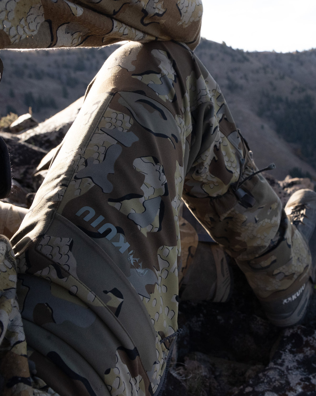 Side of Hunter Sitting on Glassing Knob Wearing Attack Pants in Valo Camouflage Highlighting YKK Zippered Cargo Pocket