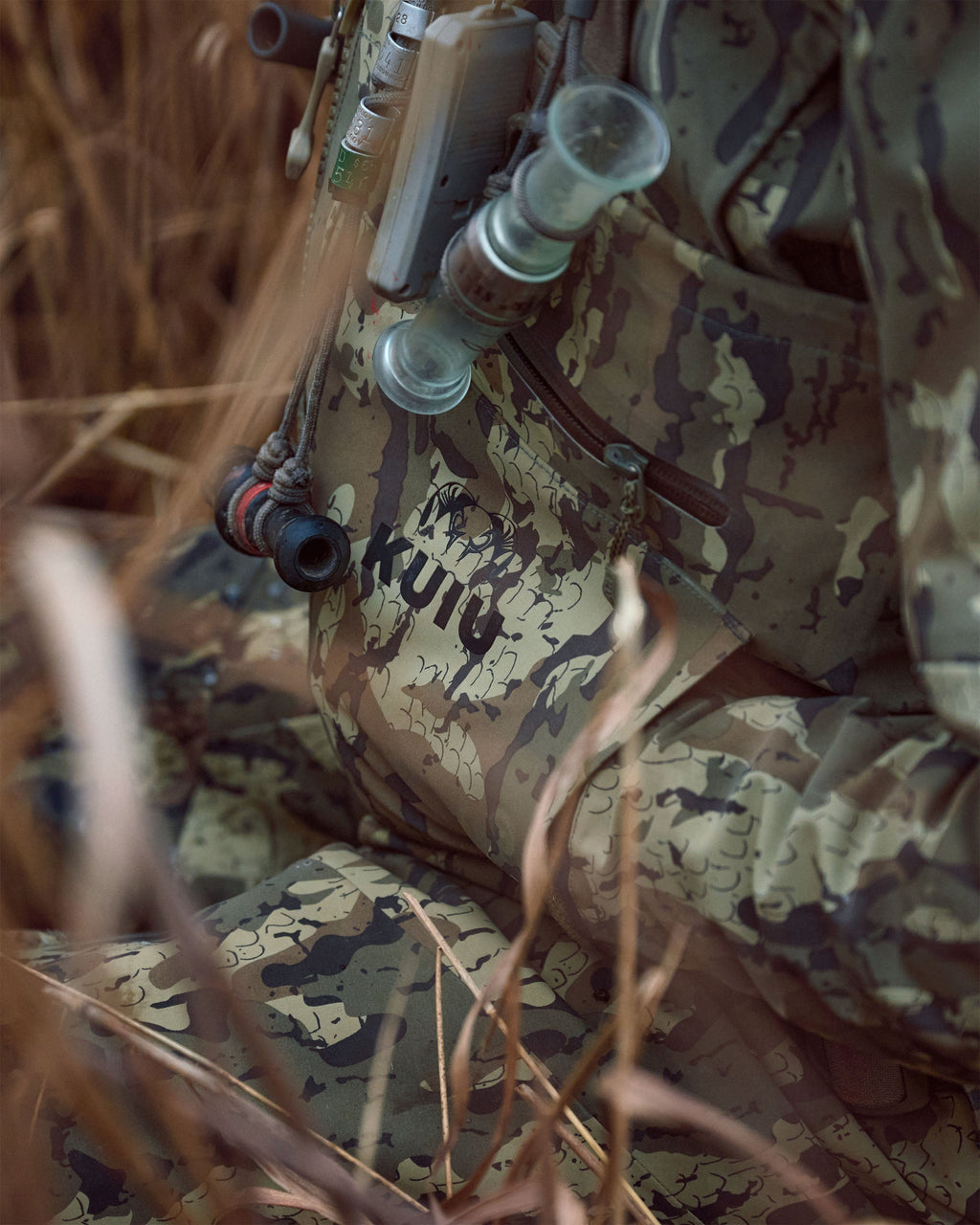 Close-up of a person in KUIU PRO HD Flex Zip Wader in Veros camouflage with a lathe tool attached, surrounded by dry grass in an outdoor setting.