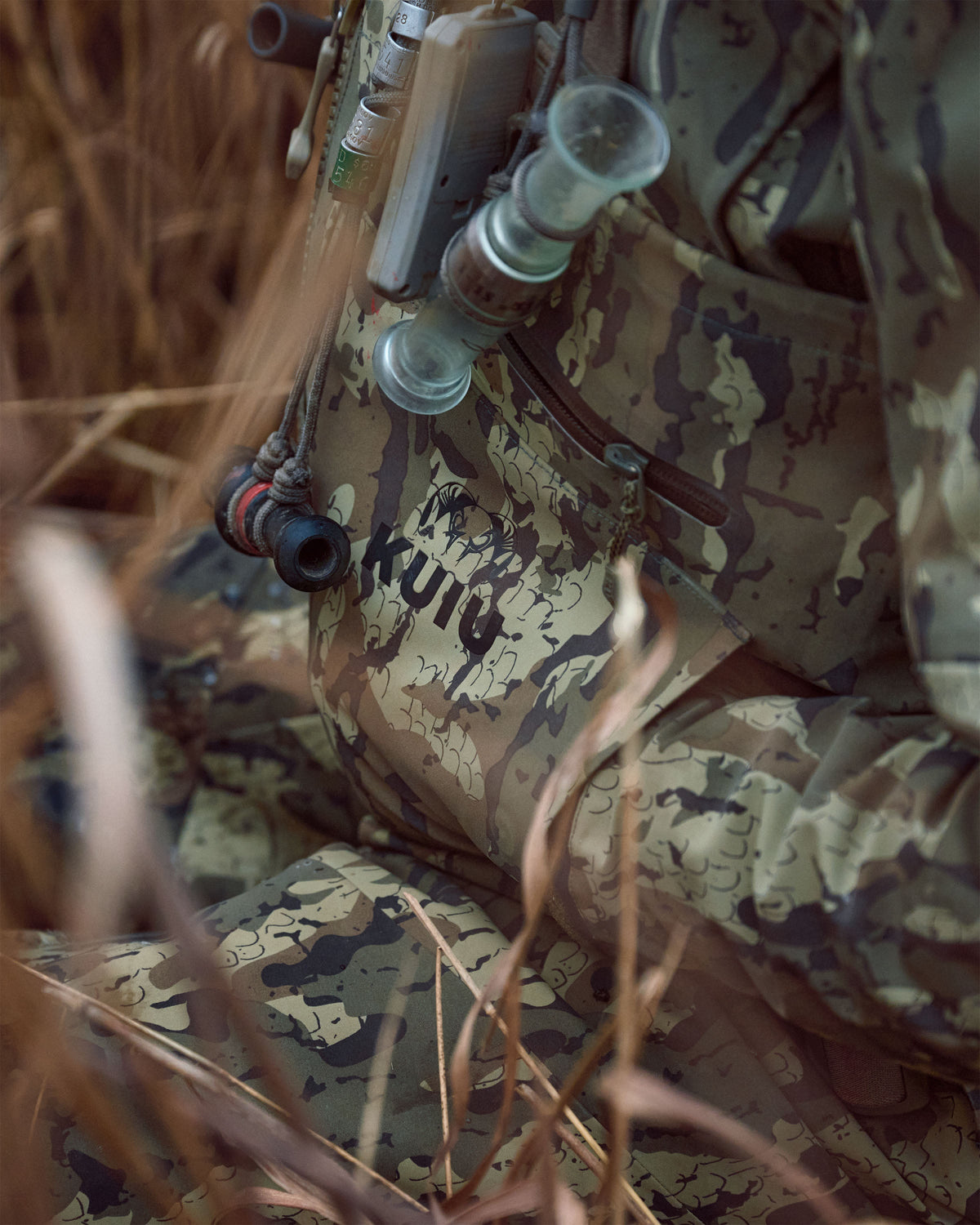 Close-up of a person in KUIU PRO HD Flex Zip Wader in Veros camouflage with a lathe tool attached, surrounded by dry grass in an outdoor setting.