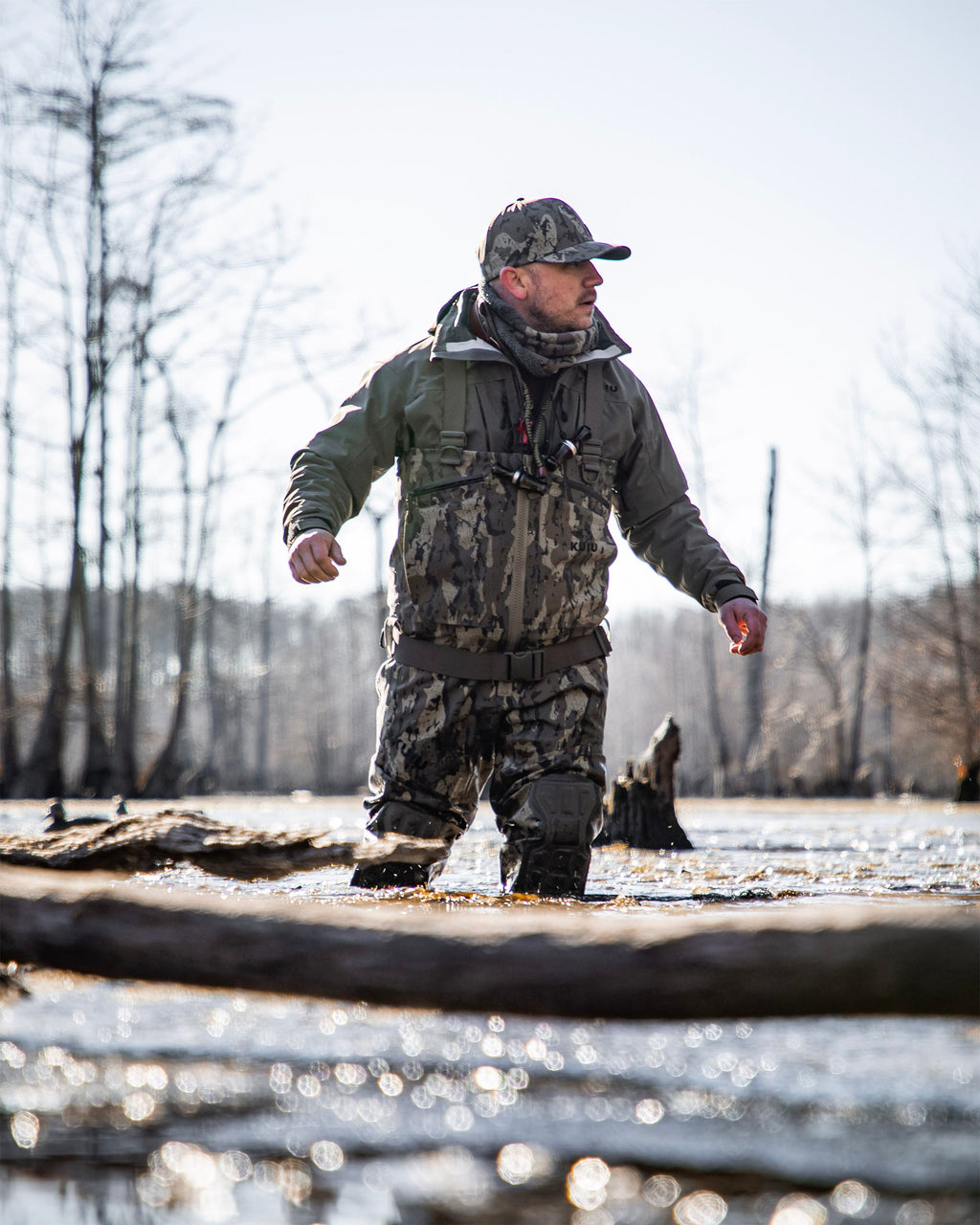 Waterfowl hunter wearing KUIU PRO HD Flex Zip Wader in Veros camouflage, standing in shallow water during a winter day with bare trees in the background.
