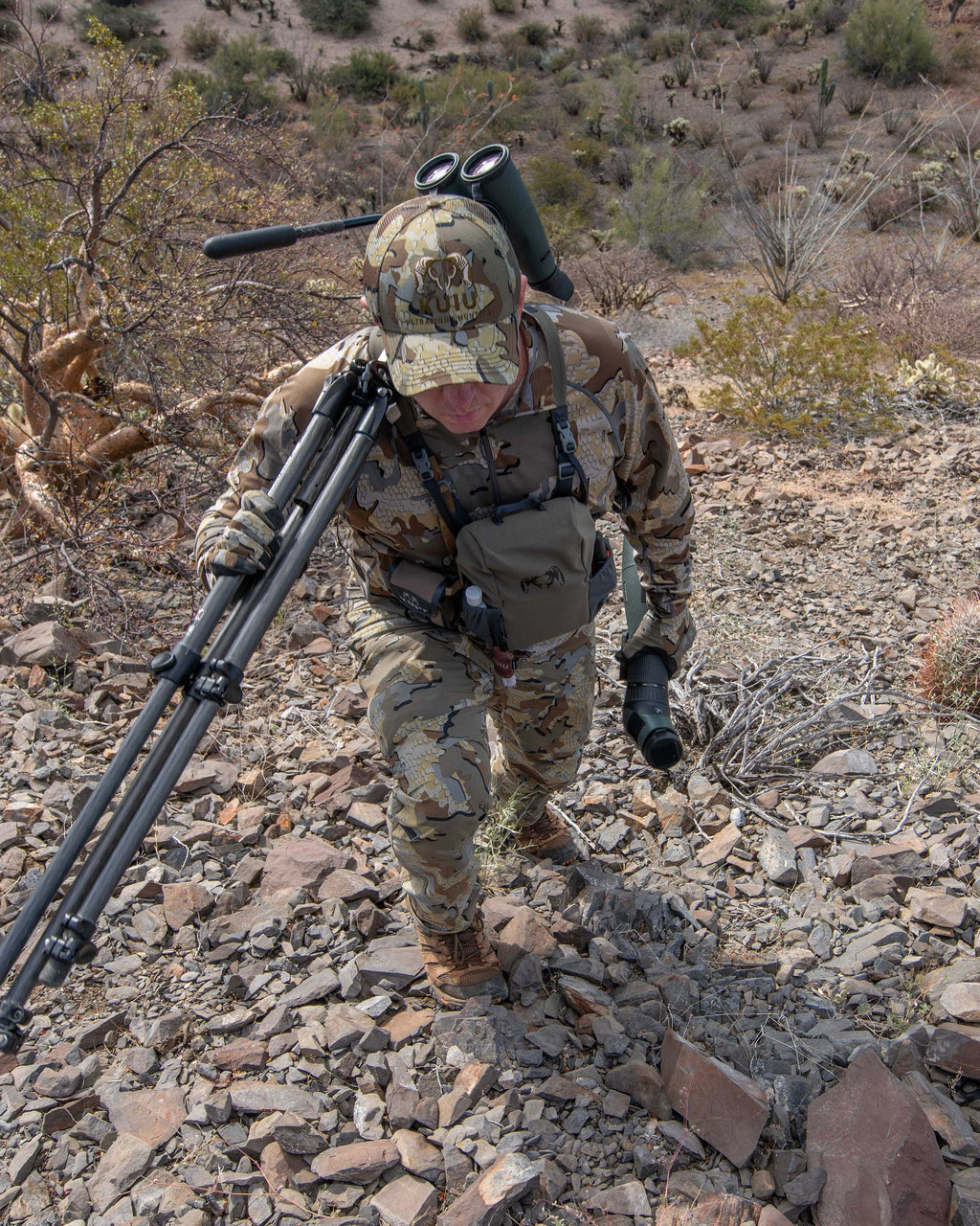 Hunter hiking up a rocky slope, carrying a spotting scope and tripod over his shoulder, wearing Kutana Stretch Woven Pant in Valo Camouflage