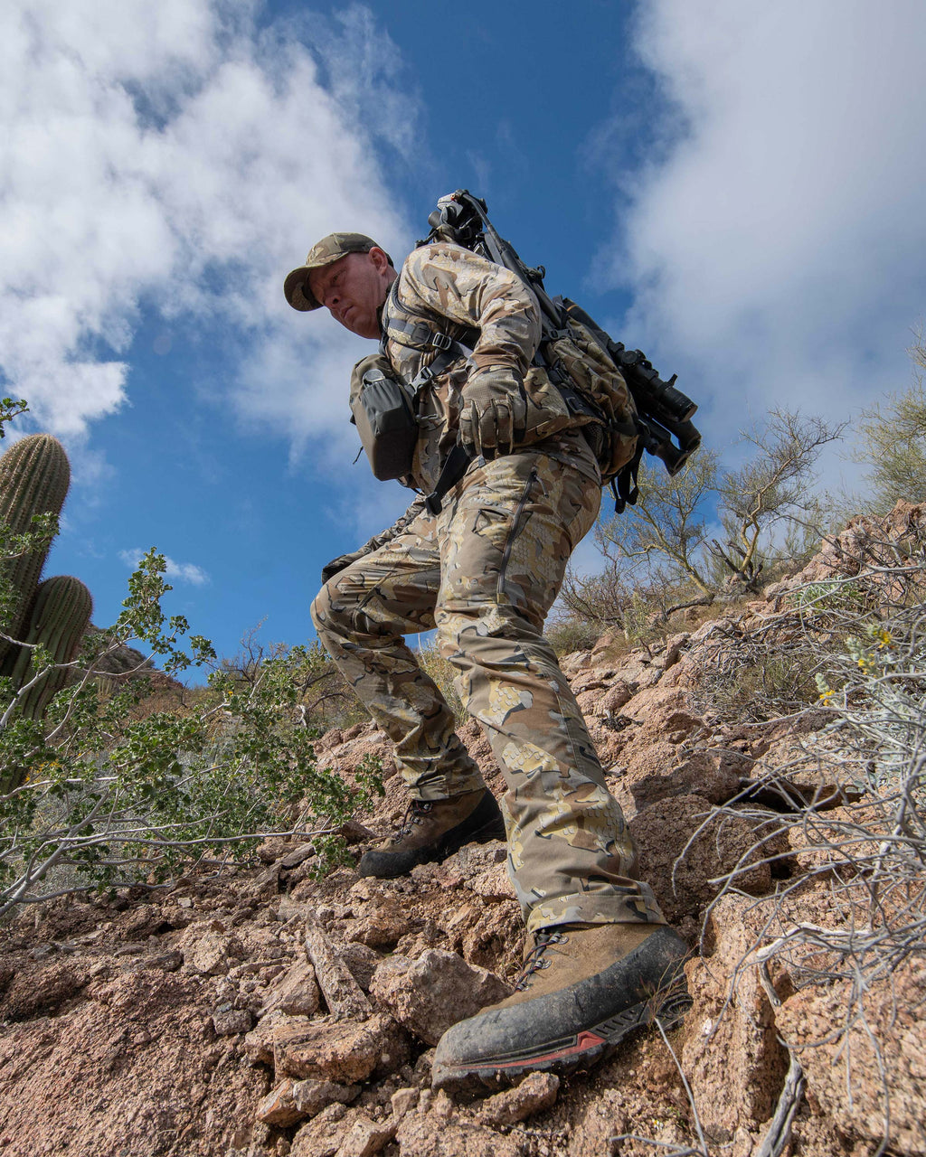 Hunter wearing Kutana Stretch Woven Pant in Valo Camouflage, carefully hiking down a rocky slope