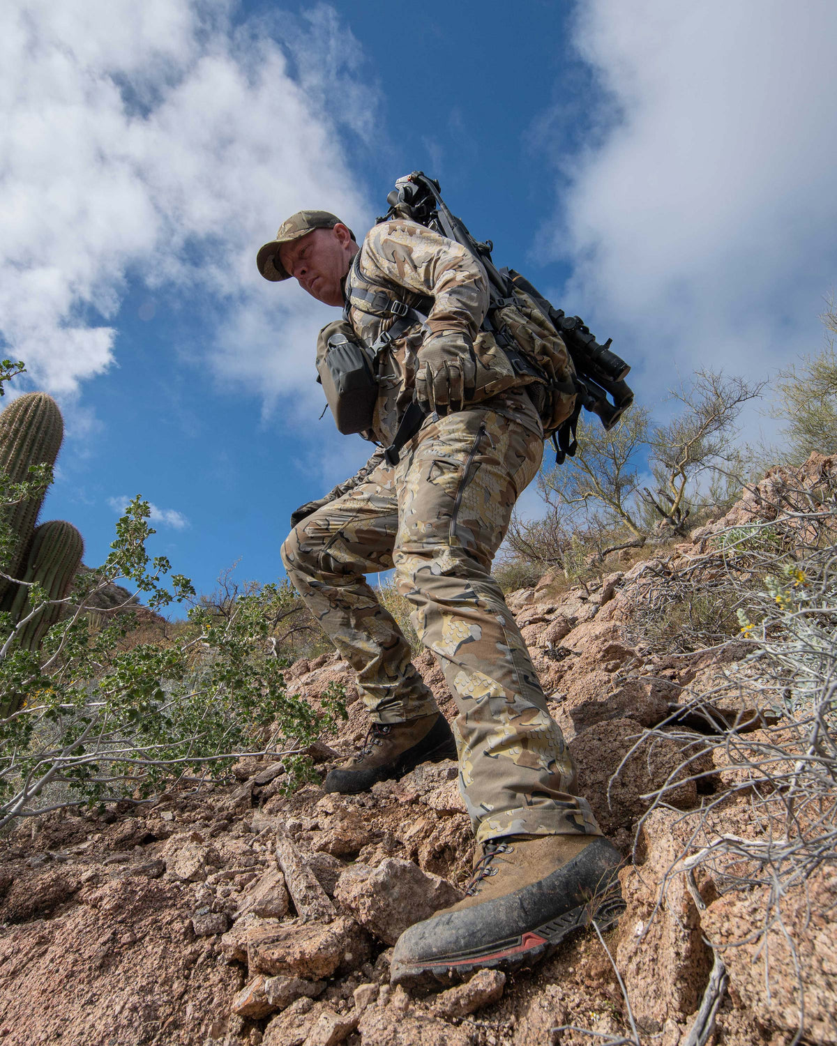 Hunter wearing Kutana Stretch Woven Pant in Valo Camouflage, carefully hiking down a rocky slope