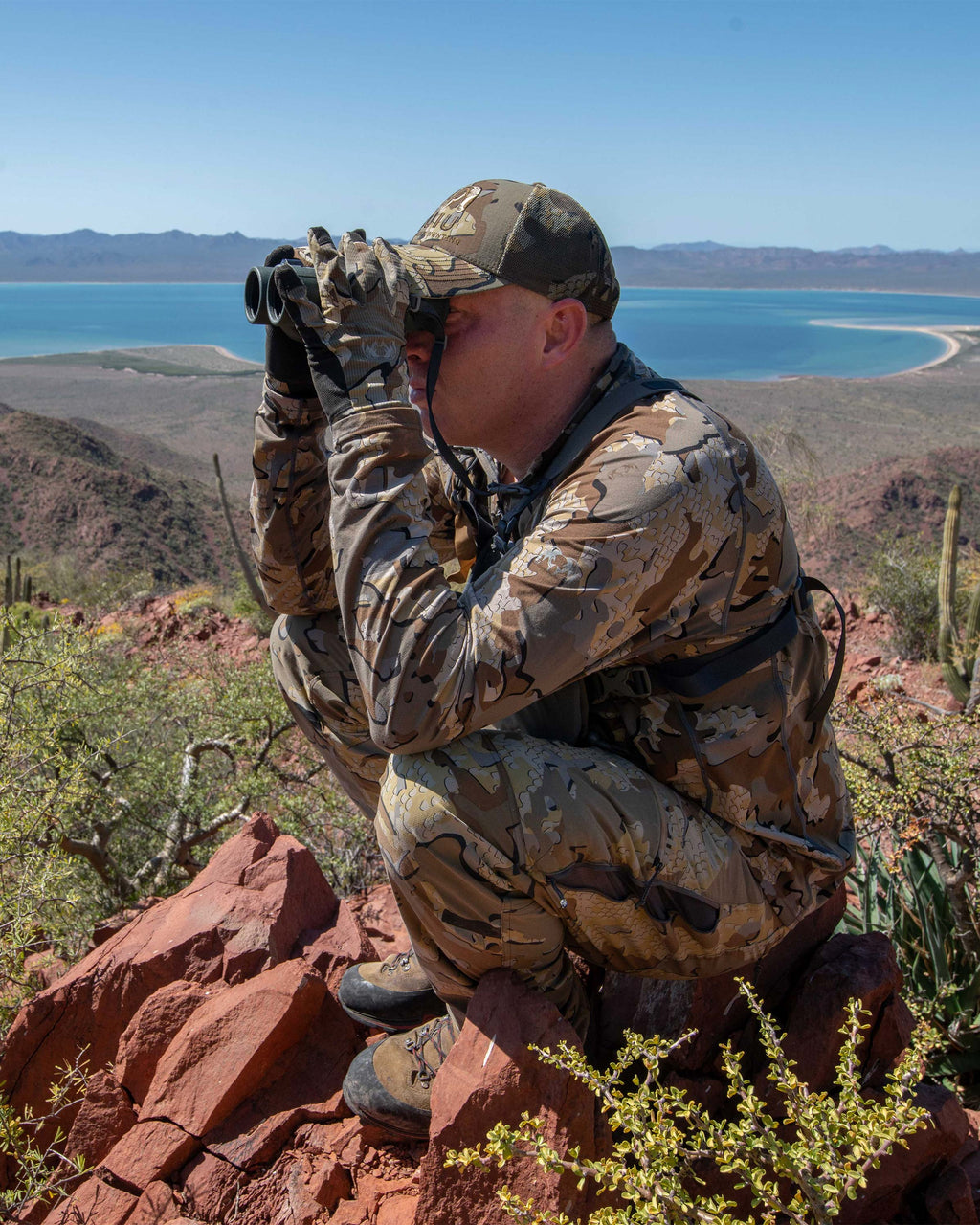 Hunter squatting on a vista point glassing, wearing Kutana Stretch Woven Pant in Valo Camouflage